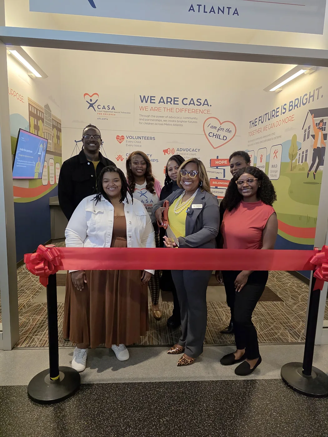 Seven people stand behind a red ribbon in front of the Atlanta CASA storefront, one holding a pair of large scissors, with a wall reading We Are CASA behind them