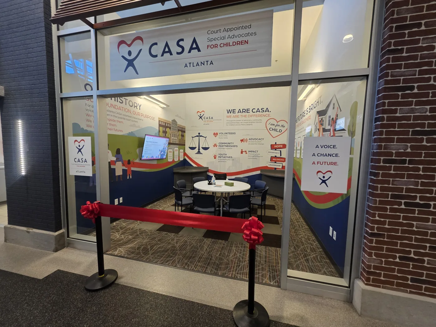 The empty Atlanta CASA storefront with a round meeting table, chairs and a wall screen, behind a red ribbon on stanchions