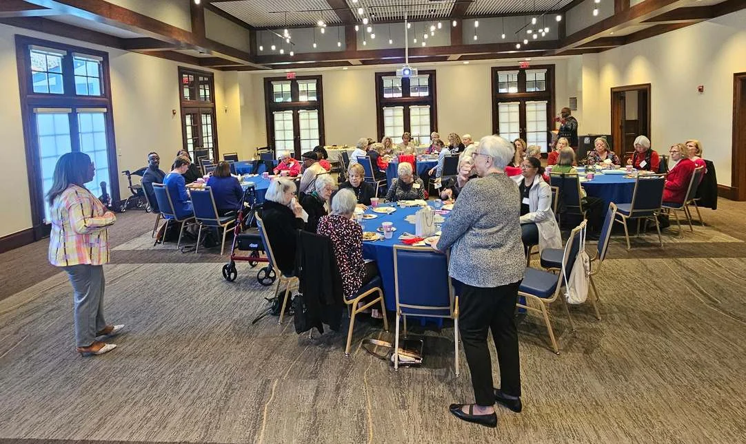 Dozens of people seated at round tables with blue tablecloths in a large hall, several standing, listening to a speaker