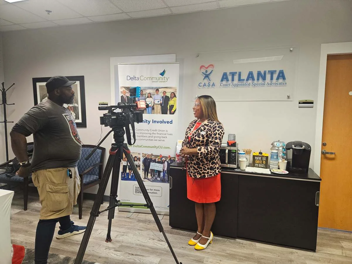 A camera operator films a woman speaking in the Atlanta CASA office, beside a Delta Community banner